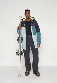 Skijas in blauw, grijs en zwart met een felgele binnenkant, gecombineerd met zwarte skibroek. Model houdt ski's vast en draagt skischoenen.