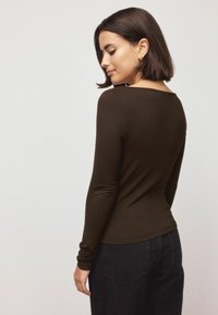 Brown long-sleeve shirt with a fitted design and a scooped back neckline, made of soft, smooth material. Paired with dark jeans.