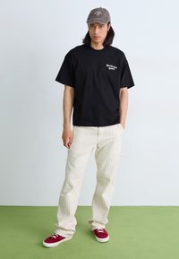 T-shirt en coton noir avec un logo blanc, pantalon rayé beige et baskets rouges avec des lacets blancs. Une casquette grise complète la tenue.