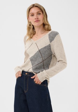 Mujer rubia con suéter de rombos beige y gris, vaqueros azules, pendientes de aro dorados y collares en capas, posando con las manos en los bolsillos.