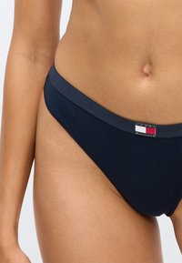 Tamni plavi bikini donji dio od pamuka s glatkom teksturom, s čvrstim pojasom i malom oznakom s logotipom uz crvene, bijele i plave akcentne detalje.