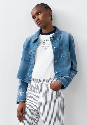 Νεαρός άτομο που φορά cropped τζιν μπουφάν με κεντήματα λουλουδιών, λευκό πουκάμισο με σχέδιο άγκυρας και παντελόνι με κάθετες ρίγες, στέκεται μπροστά σε λευκό τοίχο.