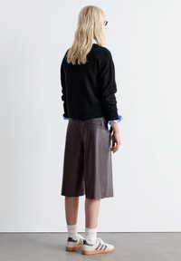 Braune Ledershorts mit einem lockeren Schnitt, kombiniert mit einem schwarzen Pullover, hellblauen Hemdärmeln und weißen Sneakers mit braunen Streifen.