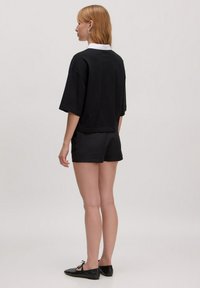 Zwarte top met korte mouwen en witte kraag, gecombineerd met zwarte shorts. Het materiaal lijkt glad, en het ontwerp is ontspannen, minimalistisch.