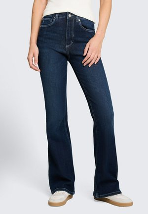 Kobieta nosząca ciemnoniebieskie jeansy typu bootcut z przednimi kieszeniami i białe trampki, stojąca z jedną ręką opartą na udzie.