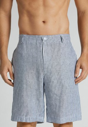 Man draagt blauw-witte kortebroek met verticale strepen en een hoge taille met knoopsluiting, handen rusten langs het lichaam, blote buik zichtbaar.