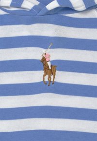 Blå och vit randig tyg med en broderad polospelare på en brun häst. Spelaren bär en rosa skjorta och håller en klubba.