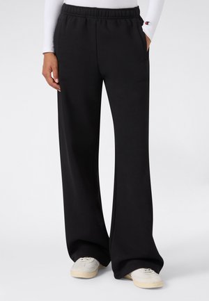 Persona che indossa pantaloni della tuta neri a gamba larga con elastico in vita e sneakers bianche, in piedi con una mano in tasca.