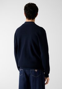 Maglione lavorato a maglia di colore blu navy con collo alto, trama a coste e maniche lunghe, abbinato a pantaloni in denim scuro con un logo triangolare sulla tasca posteriore.
