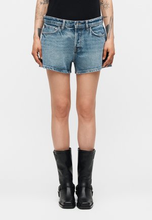 Άτομο που φοράει ξεθωριασμένα μπλε τζιν σορτς, μαύρα δερμάτινα μποτάκια μέχρι τη μέση της γάμπας και μαύρο τοπ, στέκεται μπροστά σε απλό φόντο.