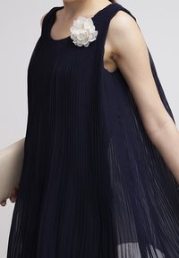 Navy sinimuotoinen hame, jossa on hihattomat käsivarret ja valkoinen kangaskukkaaksentti olkapäällä, yhdistettynä beigeen käsilaukkuun.