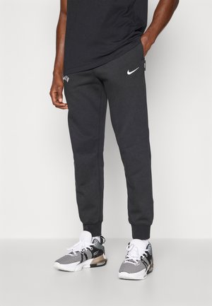 Graue Nike-Sweatpants mit Rippbündchen, weißem Logo auf dem Oberschenkel, verstellbarem Kordelzugbund, getragen mit schwarz-weißen Sportschuhen. Lässiger Fit.