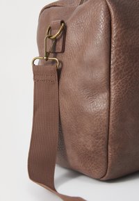 Brun kunstlædertaske med en struktureret overflade, der har en bronzefarvet metalclip og en vævet stofrem. Kasseformet, robust design.