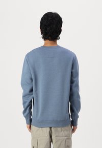 Kék pulóver kerek nyakkal, bordás mandzsettákkal és szegéllyel. Oldalt egy kis címke található. Puha anyagból készült. Minimalista design.
