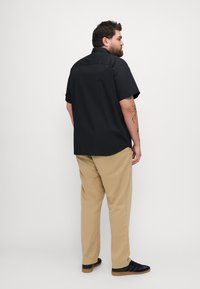 Korte mouw zwarte shirt, standaard kraag, rechte zoom, gecombineerd met kaki broeken en zwarte sneakers. Gezien vanuit een zij- en achteraanzicht.