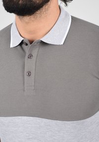 Polo gris avec un bas contrastant en gris clair. Il présente un tissu texturé, un col à revers et deux boutons marron. Rehaussé d'un liseré blanc sur le col.