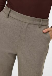 Primo piano di una persona che indossa un top marrone e pantaloni beige a vita alta con passanti per cintura, con una mano infilata nella tasca destra.