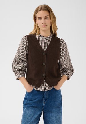 Femme aux cheveux blonds porte un gilet marron boutonné sur une chemise à carreaux et un jean bleu, debout les mains dans les poches devant un fond uni.