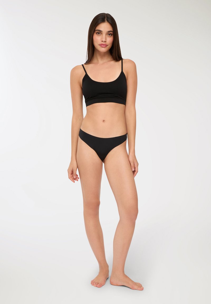 Reggiseno sportivo nero con spalline sottili e bikini coordinato, caratterizzato da una texture liscia e un design semplice. Modella in piedi a piedi nudi con capelli lisci.