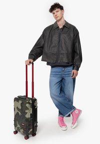 Černá kožená střihová bunda, volné modré džíny, růžové tenisky Converse a zelený maskáčový kufr na kolečkách s červenými prvky.