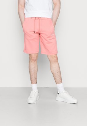 Mężczyzna w koralowo-różowych krótkich spodenkach z sznurkiem, białych trampkach, białych skarpetkach i białej koszulce stoi na jednolitym tle.