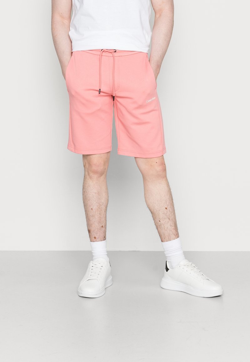 Homme portant un short corail avec cordon de serrage, des baskets blanches, des chaussettes blanches et une chemise blanche, se tenant devant un fond uni.