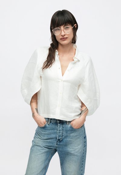 Λευκό πουκάμισο από λινό με κουμπιά και φουσκωτά μανίκια, συνδυασμένο με ανοιχτό μπλε τζιν τζιν. Το ύφασμα έχει υφή, προσδίδοντας μια χαλαρή εφαρμογή.