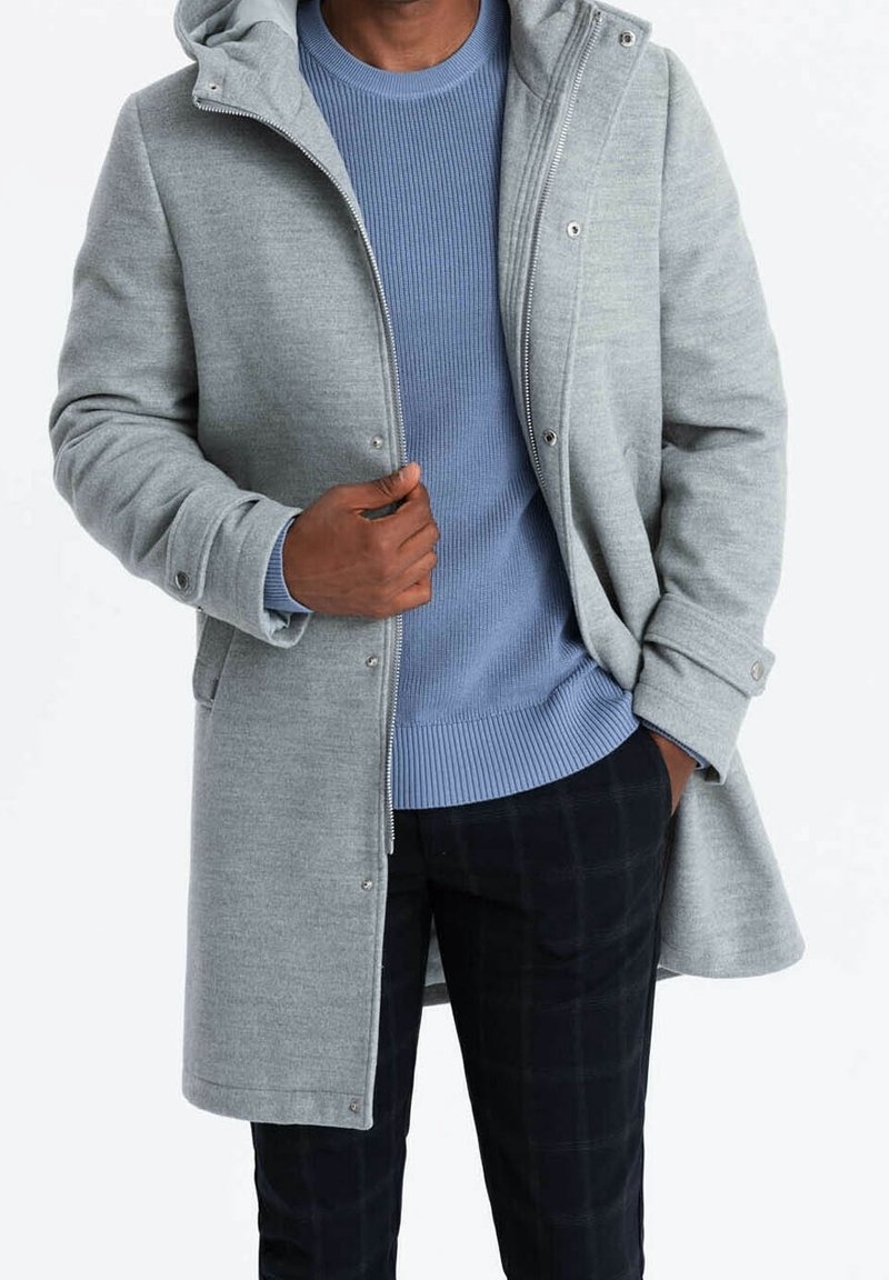 Abrigo gris de mezcla de lana con capucha incorporada, cierre frontal de broches y dos bolsillos laterales. Debajo, se ve un suéter de punto acanalado azul claro.