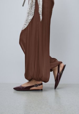 Gros plan d'une personne portant un pantalon marron ample et des ballerines slingback en cuir verni marron foncé, debout sur un sol gris clair.