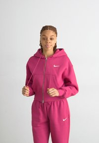 Fuchsiová mikina s kapucňou a zodpovedajúce tepláky vyrobené z mäkkej látky, s predným zipsom, klokanou kapsou a bielym logom Nike swoosh.