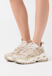 Des baskets beiges en maille et matériaux synthétiques, avec une semelle blanche épaisse et un laçage distinct avec des accents beige clair, offrant un look texturé.