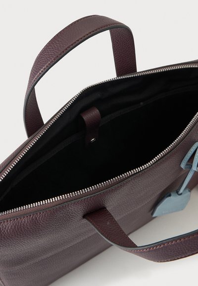Sac à main en cuir brun foncé texturé avec deux poignées, ouverture zippée en haut et une petite étiquette en cuir bleu attachée près du zip.