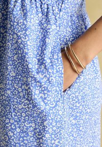 Sinine kleit valge abstraktse mustriga, millel on tasku. Kangas on kerge ja sile. Käe ümber on kuldsed käevõrud.