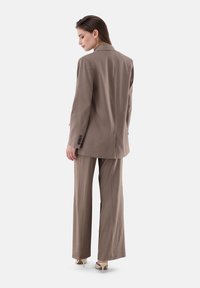 Beige maßgeschneiderter Anzug mit einem einreihigen Sakko und weit geschnittenen Hosen. Glattes Gewebe mit dezentem Glanz und Knopfdetails an den Ärmeln.