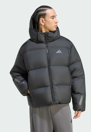 Giovane con i capelli intrecciati che indossa una giacca imbottita nera con cappuccio e pantaloni grigi, guarda di lato su uno sfondo semplice.