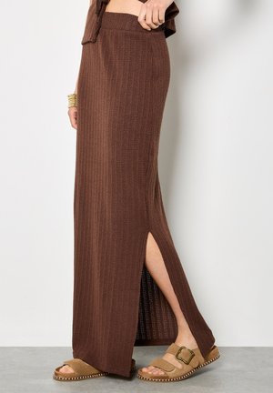 Femme portant une longue jupe en tricot marron avec une fente sur le côté et des sandales tan à boucle, debout devant un fond clair uni.