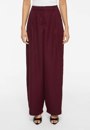 Femme portant un pantalon bordeaux taille haute à jambes larges, associé à des talons ouverts noirs et un haut blanc rentré.