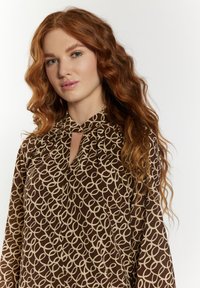 Braune Bluse mit einem beigen abstrakten Muster, Ausschnitt mit Schlitz und plissierten Details. Hergestellt aus leichtem Stoff mit fließenden Ärmeln.