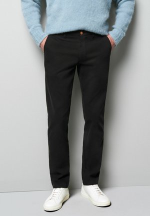 Black slim-fit trousers with a smooth texture, featuring a front button closure and side pockets, paired with white sneakers and a blue sweater.
