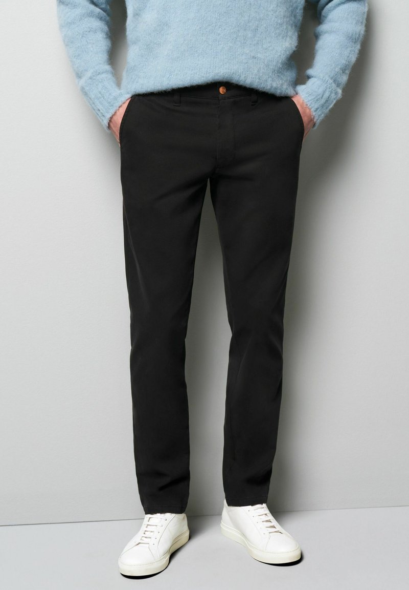 Zwarte slim-fit broek met een gladde textuur, voorzien van een sluiting met een knoop aan de voorkant en zijzakken, gecombineerd met witte sneakers en een blauwe trui.