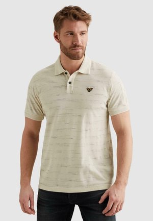 Homme portant un polo beige texturé avec un petit logo brodé sur la poitrine, assorti à un jean foncé, regardant légèrement sur le côté.