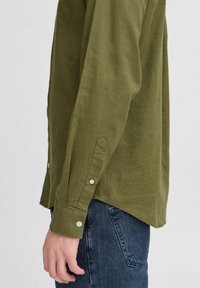 Langermet skjorte i olivengrønn, laget av strukturert stoff, med knappedetaljer ved ermene og rett kant. Kombinert med mørke denimbukser.