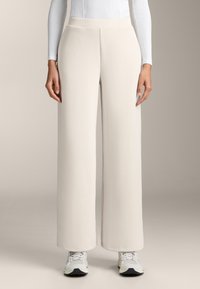 Hlače širokog kroja od svijetlo bež rebrastog materijala, s visokim strukom i glatkom teksturom. U kombinaciji s bijelim tenisicama.