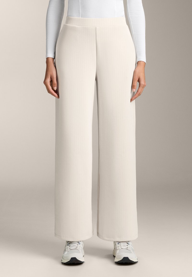 Hlače širokog kroja od svijetlo bež rebrastog materijala, s visokim strukom i glatkom teksturom. U kombinaciji s bijelim tenisicama.