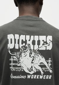 Άνδρας που φοράει σκούρο πουκάμισο με λευκό κείμενο "DICKIES" και γραφικό ενός ταύρου που πηδάει πάνω από φράχτη, μαζί με το κείμενο "genuine WORKWEAR".
