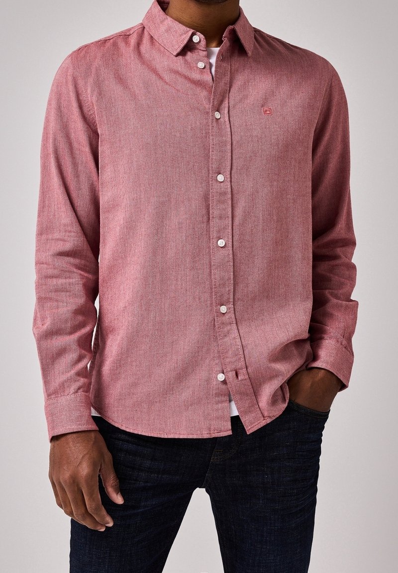 Homme portant une chemise rose à manches longues boutonnée sur un sous-vêtement blanc, jean foncé, main gauche dans la poche et main droite le long du corps.