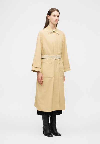 Femme portant un trench-coat beige ceinturé avec de grandes poches avant, sur une jupe plissée noire et des bottes à talons noires, debout devant un fond blanc.