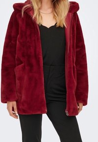 Vrouw in een dieprode, pluche hoodie over een zwarte top en een paar strakke zwarte broek, staand tegen een effen lichte achtergrond.