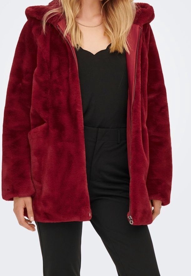 Vrouw in een dieprode, pluche hoodie over een zwarte top en een paar strakke zwarte broek, staand tegen een effen lichte achtergrond.