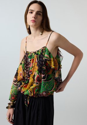 Frau trägt eine bunte, gemusterte ärmellose Bluse mit dünnen Trägern, einen schwarzen Rock, große Ohrringe und Armbänder, posiert vor einfachem Hintergrund.